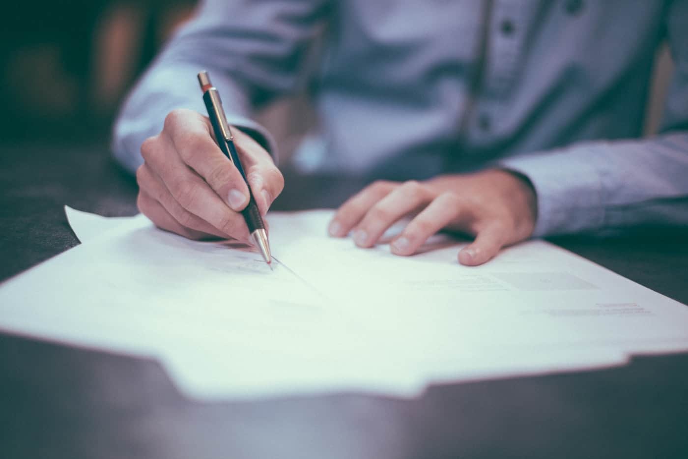 Hands signing legal documents with a pen at a dark wooden desk