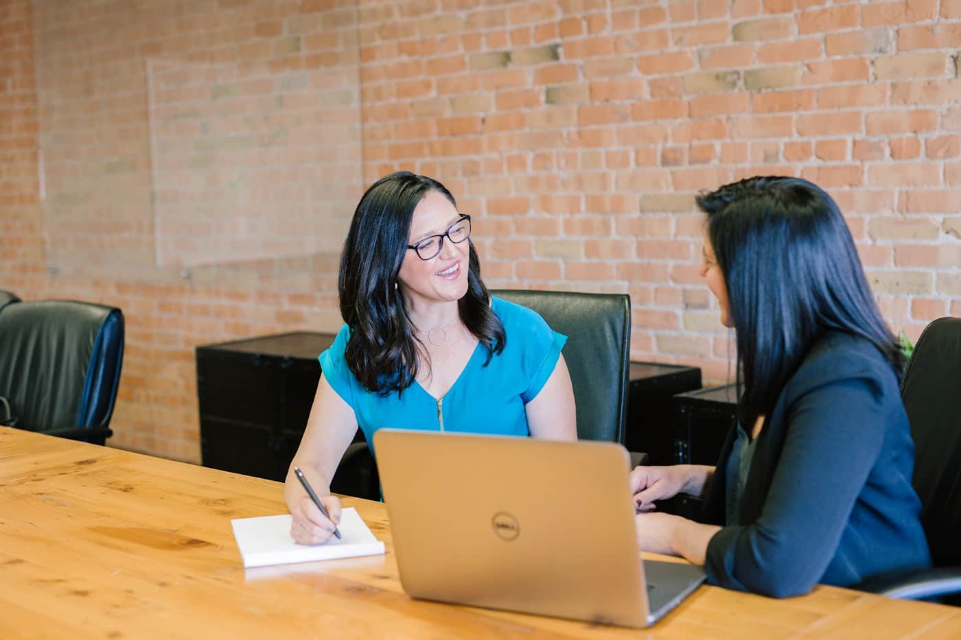 Two women in a quiet meeting at a wooden conference table, one taking notes